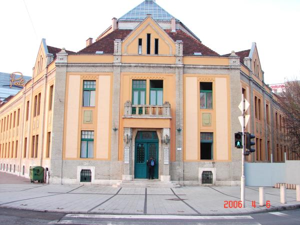 A large symmetrical multi-story building fills the frame from a street-side viewpoint. It has a peaked roof, a central entrance with a balcony above, tall windows, and pale orange wall panels separated by masonry. Sidewalk paving, traffic signs, a signal, a green bin, and a date stamp are visible in the foreground.