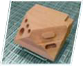 A light-brown, roughly rectangular wooden piece lies at an angle on a green grid-textured surface. Several round holes and a narrow inset slot are visible on its top face. A white angled border runs around the photograph.