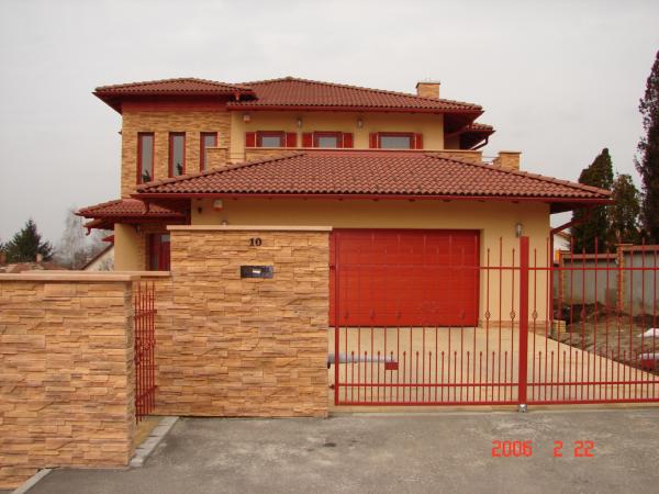 A two-story cream and tan house is seen from the street behind a red metal gate. A broad stone-faced pillar and low matching wall flank a wide red garage door, while red roof tiles cover the house. A dark number plaque is mounted on the stonework.