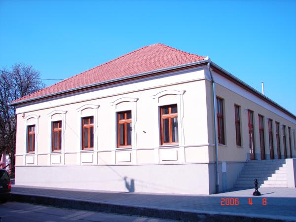 A low, light-colored building with a red tiled roof is photographed from its front corner. Tall narrow windows with reddish-brown frames run along both visible facades, each set within decorative wall molding. A broad staircase and street-side shadows appear beside the building, and a date stamp is visible in the image.