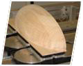 A pale wooden panel with a broad rounded top is shown leaning over a dark circular surface. A smaller curved wooden piece crosses part of the panel, and parts of dark machinery are visible beneath it. A white angled border frames the image.
